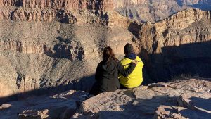 A couple sits on the edge of the Grand Canyon, overlooking the vast, layered rock formations in warm sunlight.