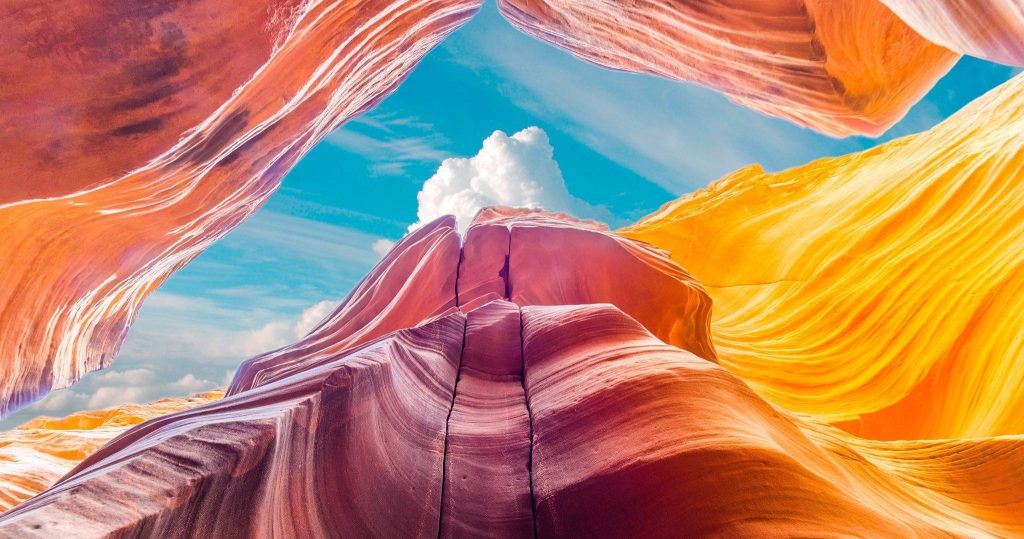 Vibrant view of a slot canyon with swirling orange and purple rock formations against a blue sky with clouds.