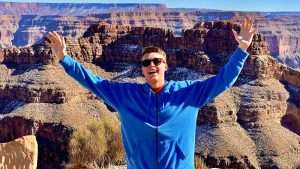 A person in a blue hoodie stands with arms raised, overlooking a stunning view of the Grand Canyon's layered rock formations.