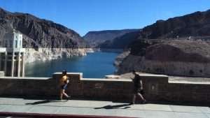 A scenic view of a blue lake surrounded by rocky mountains, with two people walking along a stone path under a clear blue sky.